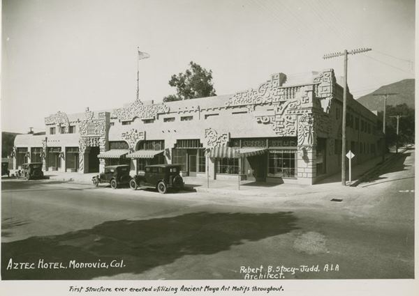 black and white 1924-5 facade of a hotel with ancient Maya art motifs, 2 stories on a corner with cars parked on street