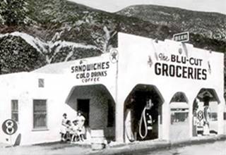 Blu-Cut groceries, US66 black and white 1940s groceries, cafe and gas station, gas pumps under canopy with arches, people to the left
