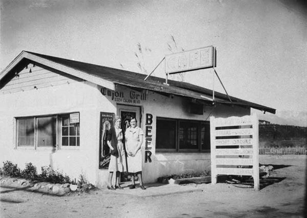 Cajon Grill US 66 San Bernardino c.1950 black and white, two women by the entrance into a gable roof, woodframe building with signs reading CAJON GRILL, BEER, CAFE