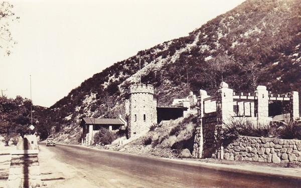 1928 Camp Cajon and US 66 sepia, highway with car stone buildings to the right on the slope, trees and stone wall and monument to the left