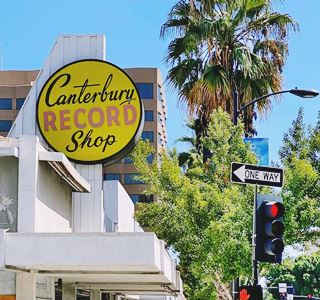 circular yellow sign in a protrusion above roofline of store reads CANTERBURY RECORD SHOP, traffic light and palm tree to the right