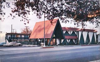 color postcard early 1960s. Steep roofed canopy over entrance, motel with units at the back 2 stories. Cars parked left. Route 66 in front. Triangles painted on wall to the right