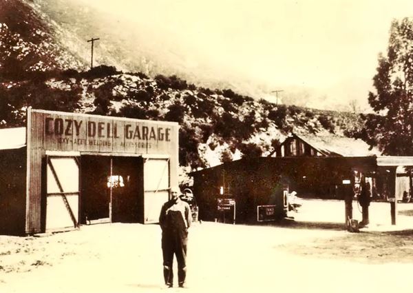 1920s Cozy Dell garage sepia, man by wood barn-like garage, small gas station with flat canopy over single pump to the left, home beyond, mountains behind. 1920s
