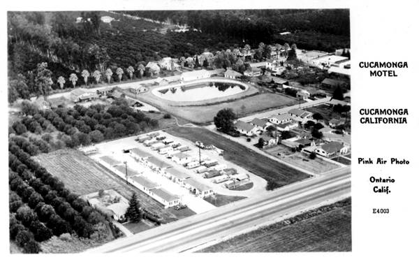 black and white 1940s photo postcard of motel seen from the air, homes and trees, US 66 runs across lower right side