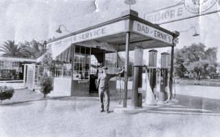 black and white, man stands by small box shaped woodframe building, flat canopy over 3 gas pumps sign reads GILMORE, canopy reads Dad’s and Ernie’s Super Service. 1939