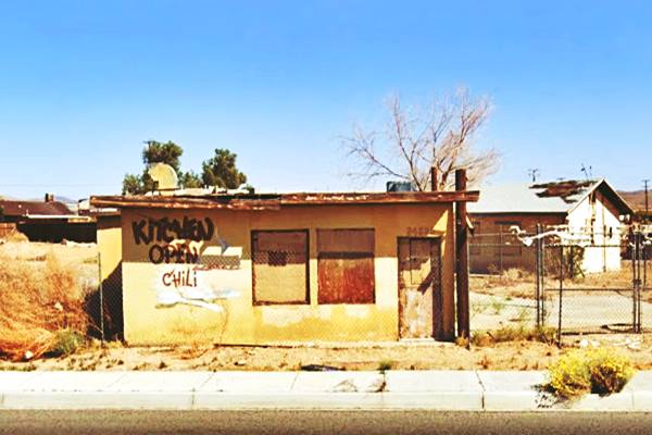 vacant decrepit box shaped, flat roof tiny cafe, KITCHEN OPEN written on the wall by the boarded windows and door. Motel behind, chain link fence in front