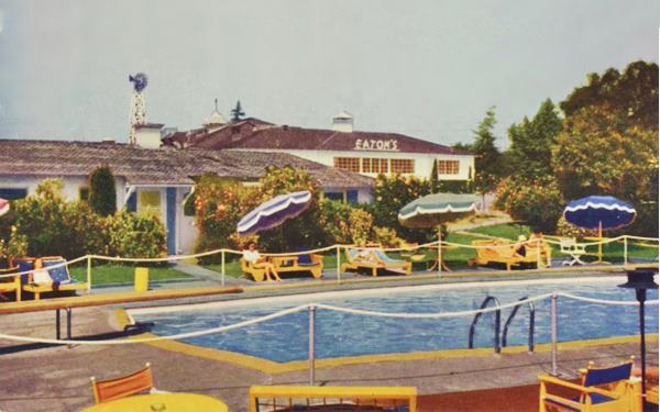 Eaton’s 1950s postcard Arcadia CA Route 66 pool scene, people on deck-chairs by pool, building behind reads EATONs on sign, trees, lawn, sunny day c.1950s