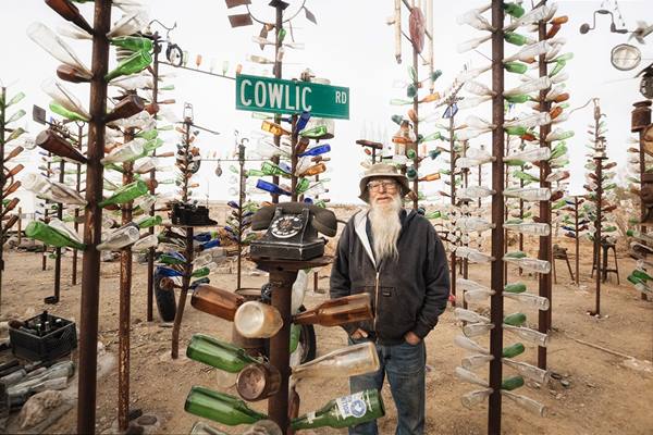 Elmer Long at his Bottle Ranch US 66 man, with white beard by large telephone artwork bottles on steel trees