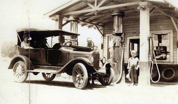 sepia, 1920s, car by pumps and gable roof canopy woodframe gas station, man pumping gas, boy beside car, 3 people (one a woman) in the car