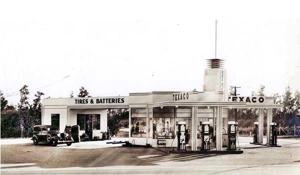 sepia, 1930s, car by gas station, flat canopy tower over office, pumps facing both streets at the corner, tires and batteries annex at the rear TEXACO signage