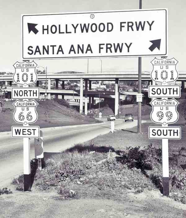 Four Level Interchange 1950s Los Angeles CA Route 66 black and white photo, 1950s cars approaching a stacked interchange with concrete pillars road signs with shields of US 66, 99 and 101, and arrows marking Hollywood Frwy and Santa Ana Frwy