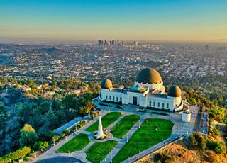 Griffith observatory Los Angeles color aerial view, skyscrapers in the distance, downtown LA, and a white stone building with a park and domes, for Planetarium and observatory on top of a hill, city below