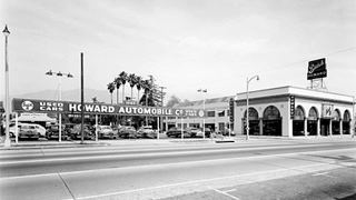 black and white 1950s auto dealer sign with cars, and on the other corner a building with arch shaped windows, sign reads HOWARD AUTOMOBILE CO. Used Cars
