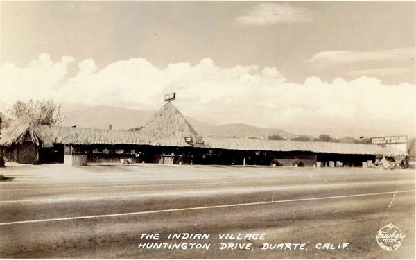 Indian Village c.1930s Duarte CA Route 66 black and white 1930s. thatched building with a conical protrusion, sign on the right, mountains beyond, Route 66 runs in front, looks like African hut, long, narrow, low