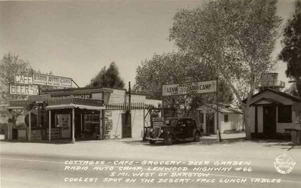 black and white 1930s postcard woodframe buildings: Cafe, Garage, car, trees, water tank beyond, US66 in front