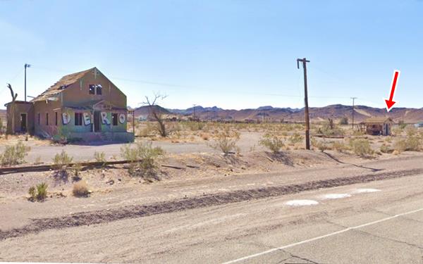 color 2021. Cottage 2 story building left vacant and decrepit, 2 cabin marked with a red arrow, dry tree trunks, hills beyond. US 66 runs in front