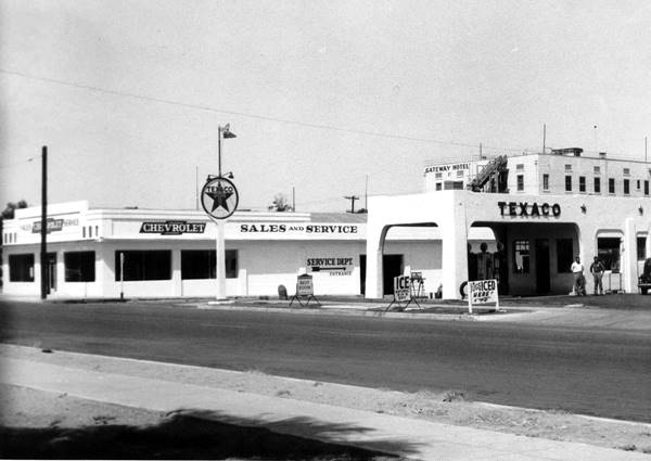black and white, Texaco gas station pueblo style (right) white box shaped Chevrolet dealer. Gateway Hotel written on building behind, several stories high c.1940
