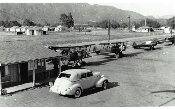 black and white c.1940 planes lined up, office with gable roof to the left and a white car, convertible beside it. 3 men under porch. Buildings beyond, a motel and a drive-in by Route 66. Mountains in the distance