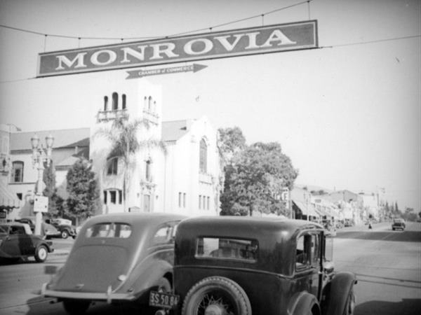 black and white early c.1937 view of Foothill looking east from Myrtle with cars and a long stretch of Foothill to the left is the Mission Revival style First Presbyterian Church, arrow says MONROVIA Chamber of Commerce hangs over road above 2 cars pointing right