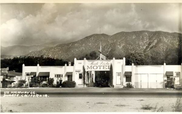 1930s Monte Vista Auto Court, Duarte CA Route 66 black and white 1930s. Single story enclosed building facing Route 66 signe reads MONTE VISTA AUTO COURT. Man and woman stand by office, mountains byond. Route 66 in front