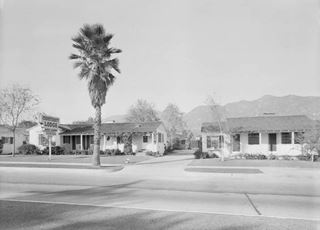 black and white, 1940s. Gable roof motel buildings with central court, neon sign and palm tree to the left, mountains behind, Route 66 in front