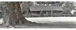My Old Kentucky Home 1940s Monrovia US66 1940s black and white photo log cabin surrounded by trees, large trunk in the foreground left. Rooftop reads MY OLD KENTUCKY HOME - DINNERS. Fence around builting and a parking area