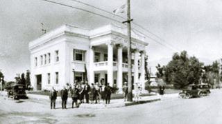 1926 Old City Hall, Arcadia black and white 1926, colonial style 2 story building with columns and US Flag, on a corner. Cars parked, men standing by it