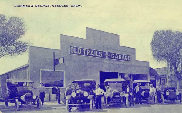 black and white view of a garage, stepped parapet, name on it: OLD TRAILS GARAGE - Lorimer and George. Five early 1920s cars and several men standing in front of it