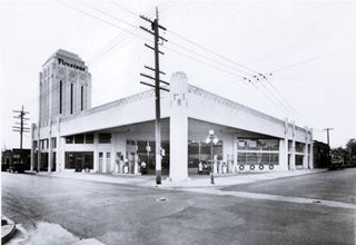 black and white, 1930s. flat roof over gas pumps, car parked, tower to the left, on a corner, a gas station. Offices and garage in the back