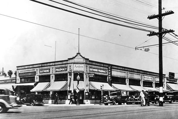 Raymond Pharmacy. South Pasadena CA Route 66 black and white 1930s, corner building, canvas awnings cars parked and driving by sign reads RAYMOND PHARMACY