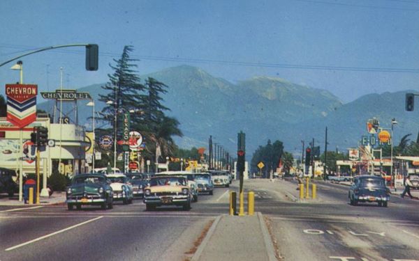 color 1950s view of cars, gas stations and neon signs looking east along Foothill Blvd at its x-ing with Riverside Ave, San Bernardino Mountains in the distance.