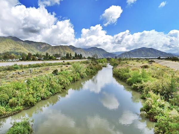 river running among bushes on its banks, mountains in the distance, coluds in the sky, a gash in the mountains marks a distant canyon