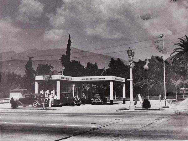 black and white 1930s, 3 cars parked under canopies of a Richfield station, mountains beyond, men, street runs in front, trees behind