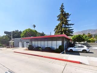 flat roof, glazed building 2 service bays and sign left, parking area right, trees behind, a former gas station, US 66 runs in front, red and gray building.
