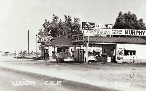 1930s black and white left US 66, right: two gas stations, one says MURPHY BROS Chevron, the other SHAMROCK CAFE, cars filling tank, gabled canopies, trees behind