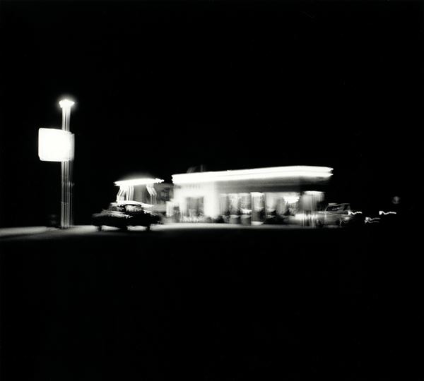 Ed Ruscha (c) 1962 From Twentysix Gasoline Stations, Shell Daggett CA black and white night photograph of a blurred lit up gas station with 2 cars 1962 Shell Daggett