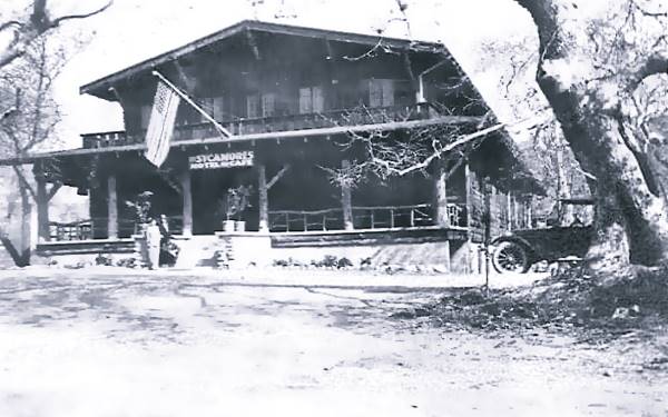 1920s black and white gabled 2 stor building HOTEL written on sigh, flag, people, trees