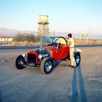 Poe Cafe c.1960s color, 1960s. Man by red sports car, Route 66 behind, and beyond a wood water tank atop a timber tower, desert shrubs, buildings in the distance and cafe sign to the right