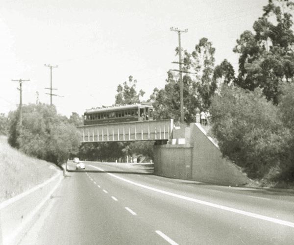 1950s black and white. Tram crossing bridge over Route 66, cars along highway. Trees line it
