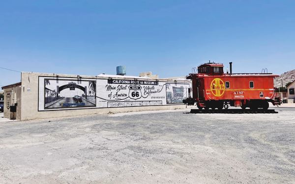 red caboose to the right, wall with black and white map-mural of US66 from Needles to Santa Monica CA, gravel parking area