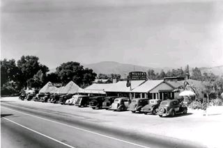 black and white c.1940s cars parked by a long gable roof building sign on roof and to the left, US66 runs in front, parasol to the left WILSONs Dinner House