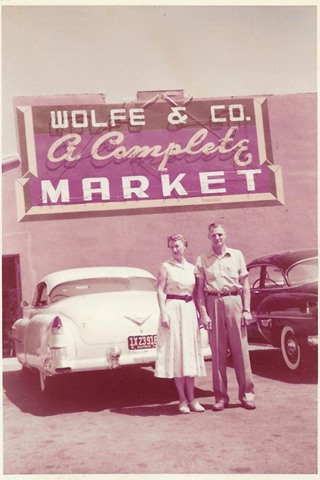 Wolfe’s Market 1950s US 66 Claremont CA color 1950s, man and woman stand by parked white car and sign above building behind them and car reads WOLFE & CO A COMPLETE MARKET, black car to the right,