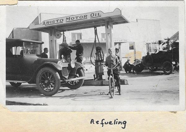 black and white, 1920s. Left: people by a car pumping gas under small roof, boy on bike, people and cars by building behind pumps