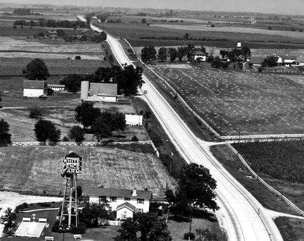 black and white c-1930s, aerial, right: US 66 runs through fields, left: building in a field, two stories, with: WHITE FENCE FARM written on the water tank beside it