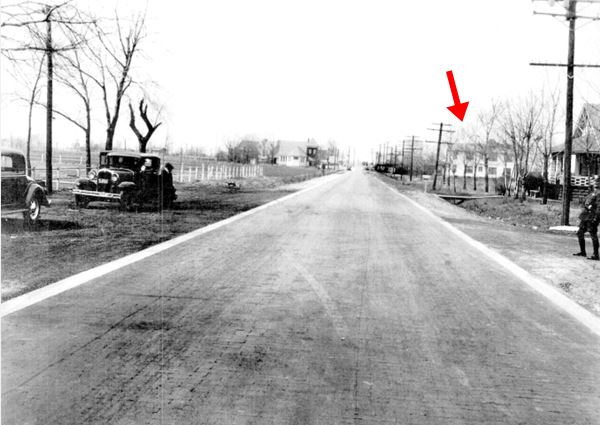 black and white photo from 1933 to the left to cars on the shoulder, a brick paved road in the middle, a policeman and houses to the right, further away more buildings