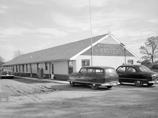 black and white, 1951, motel with gable roof long linear layout for units LEXINGTON MOTEL written on facade, 3 cars parked by it