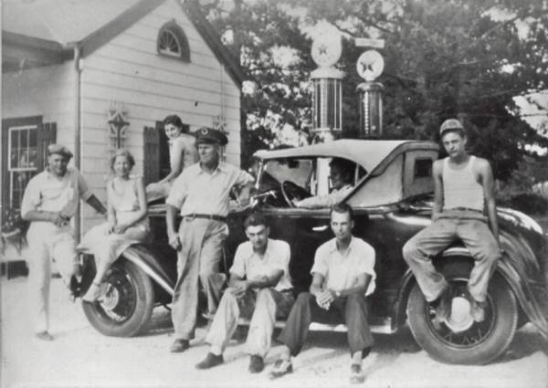 black and white c.1930s, a group of 5 men and 2 women sit or stand by a car next to a woodframe gas station with Texaco pumps