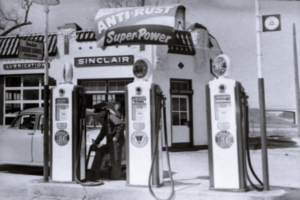 black and white c.1950s, Spanish Mission style Sinclair station, attendant filling car by pumps