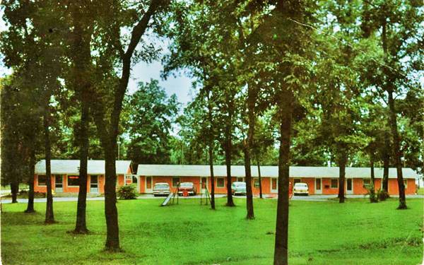 1950s view Ann Rutledge Motel color postcard, signle story long building, gable roof, painted orange-red with 1950s cars parked by the units. Trees surround it