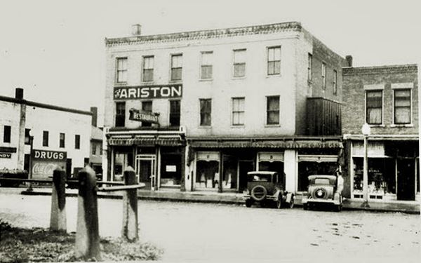 three story building on a corner with cars parked in front of it and sign "ARISTON" on facade. A black and white 1920s postcard